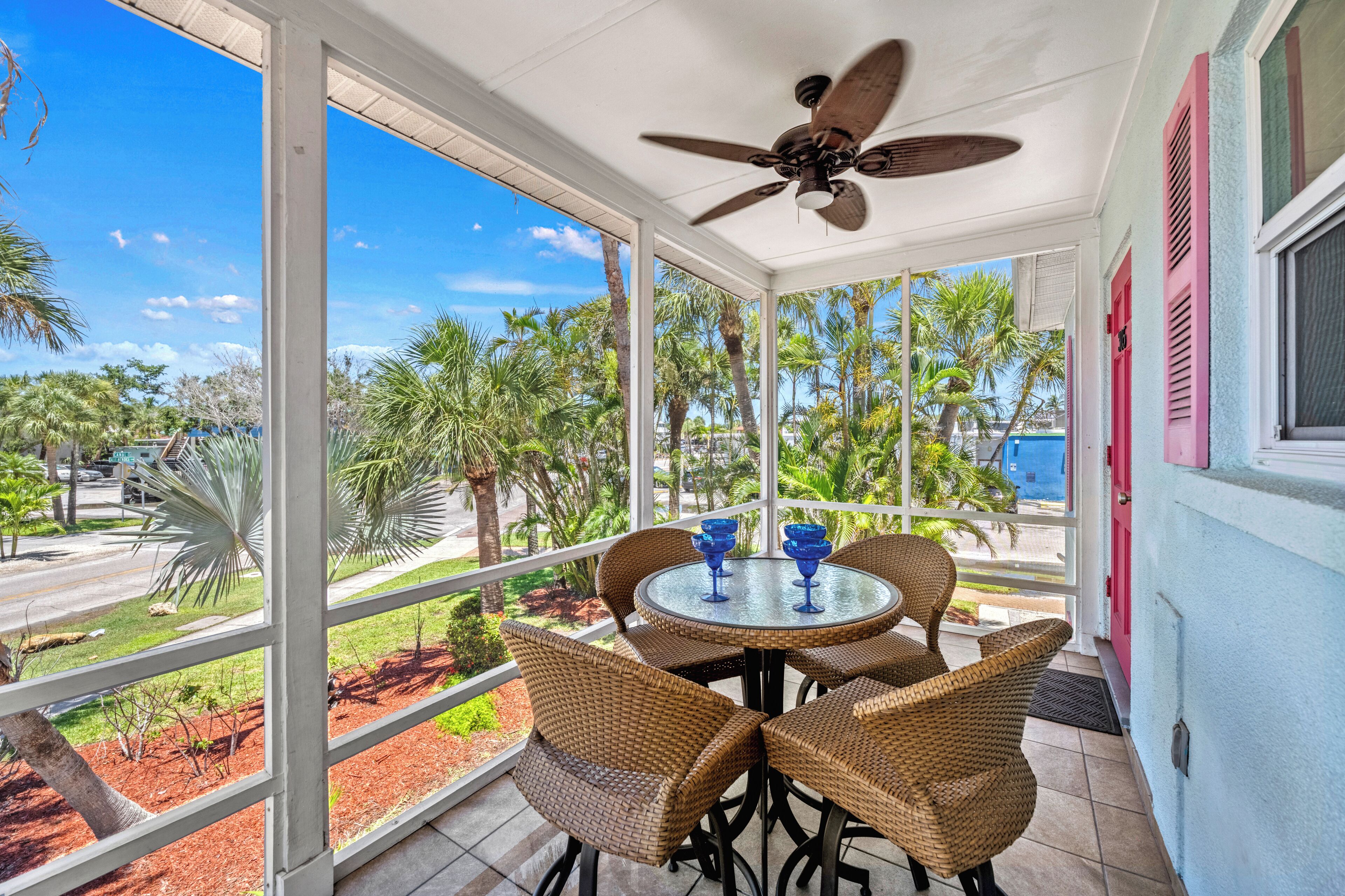 A covered outdoor area with a table and chairs, a ceiling fan, and a view of palm trees and a road.