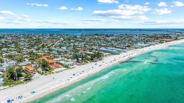 An aerial view of a coastal town with a long sandy beach, clear turquoise waters, and dense residential buildings.