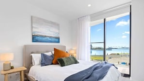 A bedroom with a large bed, a nightstand, a lamp, a painting, and a view of a body of water.