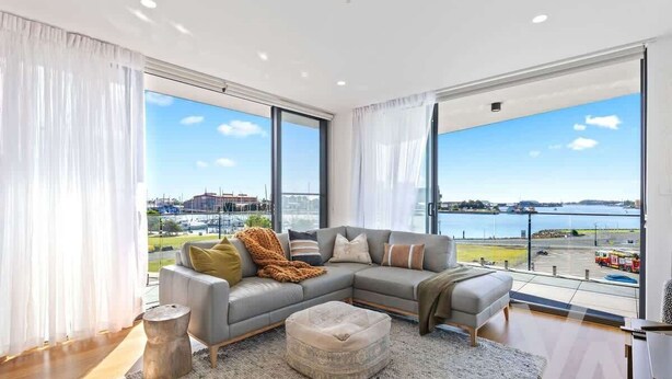 A modern living room with a grey sectional sofa, a round ottoman, and a view of a body of water and cityscape through large windows.