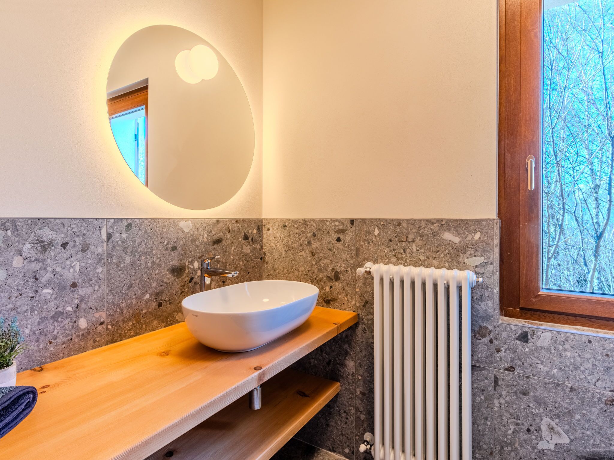 A bathroom with a white vessel sink, a wooden countertop, a stone wall, a round mirror, and a window with a view of trees.