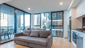 A modern apartment with a grey sofa, a kitchen with a stove and oven, and large windows offering a city view.