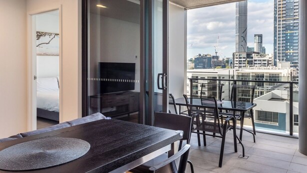 A modern balcony with a dining table and chairs, a TV in the adjacent room, and a city view.
