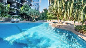 A swimming pool with clear blue water, surrounded by lounge chairs and palm trees, with modern buildings in the background.