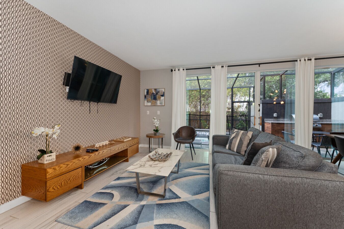 Un salon moderne avec un mur décoré de motifs, un téléviseur à écran plat, un canapé, une table basse et un coin repas équipé d’un barbecue.