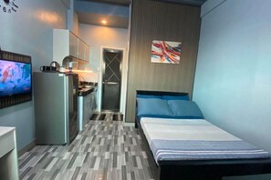 Quarto de hotel compacto com uma cama, televisão, kitchenette e porta de acesso a uma casa de banho.