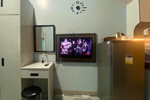 Um quarto pequeno com televisão na parede, uma escrivaninha com cadeira e um frigorífico.
