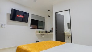 Habitación de hotel con una cama, un televisor en la pared mostrando 'NETFLIX', una ventana con persiana, un lavamanos con espejo y un ventilador de techo.