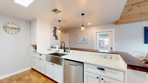 A modern kitchen with white cabinetry, a stainless steel sink, and a dishwasher. There is a wooden ceiling and a decorative wall clock. - Pet Friendly, Old Town Location, Golf Cart Access, Close to the Beach (Port Aransas)