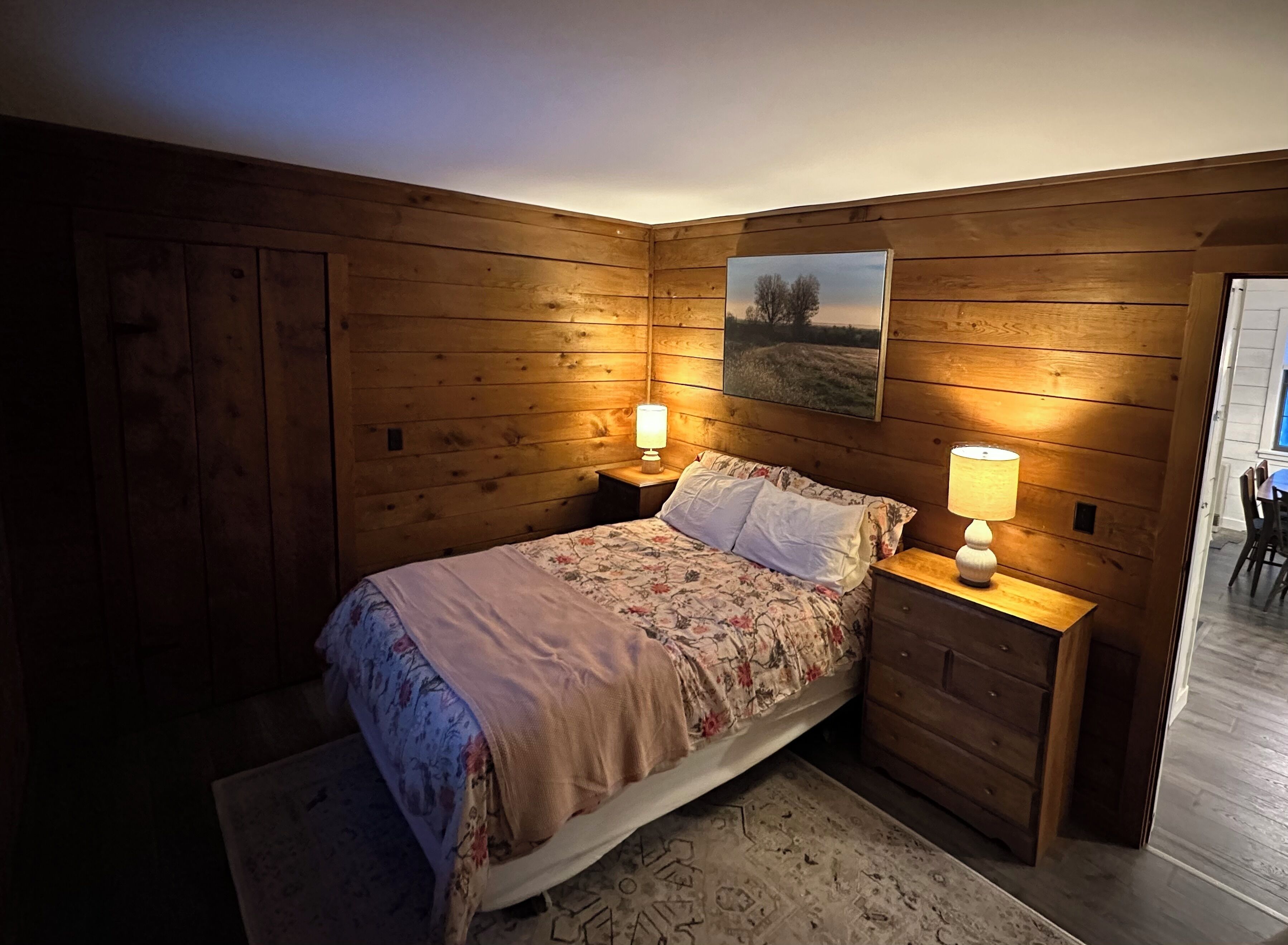 A cozy bedroom with wooden walls, a bed with floral bedding, two bedside lamps, and a painting on the wall.