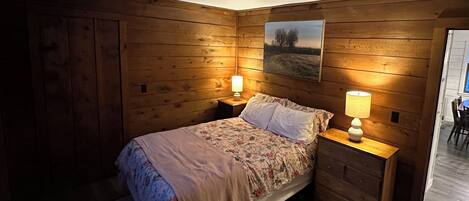 A cozy bedroom with wooden walls, a bed with floral bedding, two bedside lamps, and a painting on the wall.