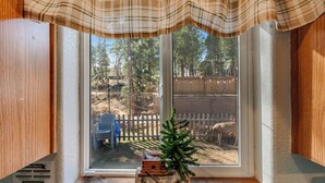 Una ventana con cortina escocesa, una pequeña mesa de madera con un árbol de Navidad, y vista a un patio cercado con árboles y una silla.