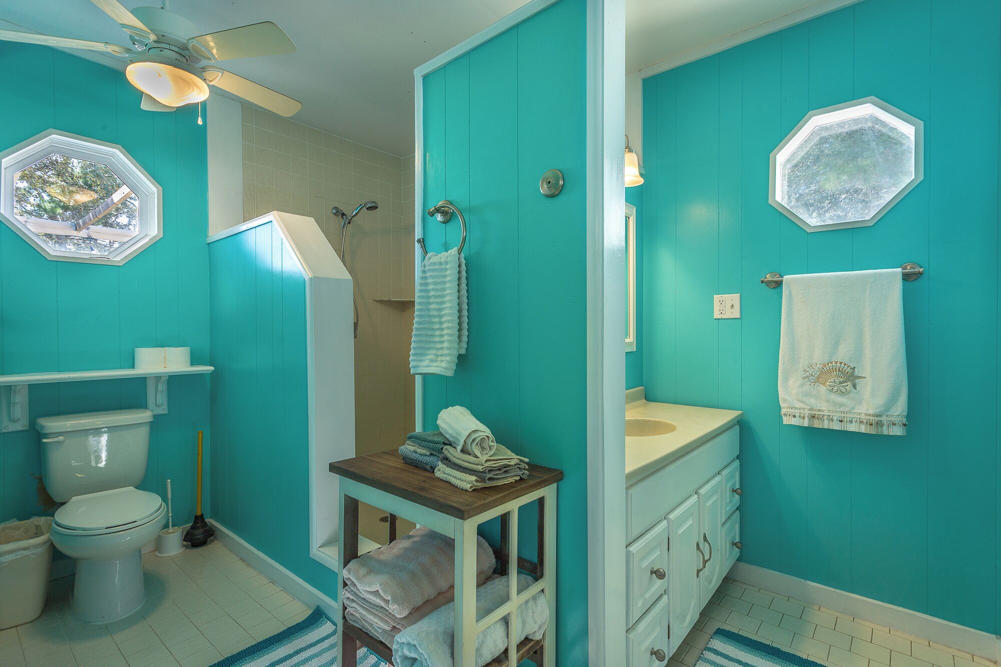 A bathroom with teal walls, a white toilet, a sink, and a shower. There are towels and a small table with books.