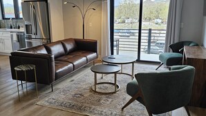 A modern living room with a leather sofa, a coffee table, and a green armchair. A ceiling fan and a large window with a view of the outside are also visible. - The Mayfly Hotel, Unit 1E (Salida)