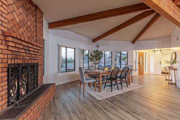 A dining area with a brick fireplace, wooden beams, and large windows.