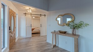 A hallway with wooden flooring, a wooden door, a bed in the adjacent room, a console table with a vase, and a mirror on the wall.