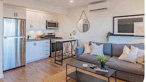 A modern kitchen with white cabinets, a stainless steel refrigerator, and a microwave. There's a round mirror on the wall, a gray sofa with cushions, and a small coffee table with a plant. - INN ON THE BENCH (Lincoln)