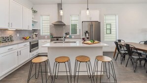 Eine moderne Küche mit weißen Schränken, einer zentralen Kochinsel mit Barhocker und einem Edelstahl-Kühlschrank, beleuchtet durch Pendelleuchten.