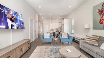 A modern living room with a flat-screen TV, a sofa, a coffee table, and a dining area with chairs.