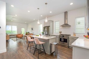 A modern kitchen with a central island, stainless steel appliances, pendant lighting, and a large window providing natural light.