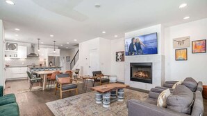 A modern living room with a fireplace, a dining area, and a kitchen in the background.