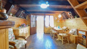 Une pièce mansardée chaleureuse avec des murs en bois et un plafond en pente. Elle comprend une table à manger en bois avec des chaises, un banc et un petit coin cuisine équipé d’une cuisinière et de placards.