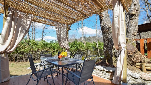 Un espace de restauration en plein air avec une table et des chaises, une pergola au-dessus et une vue sur les arbres et la verdure.