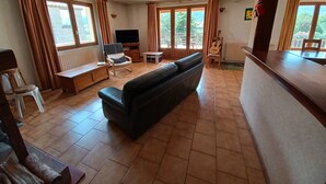 Una sala de estar con un sofá negro, muebles de madera, un televisor y ventanas grandes con cortinas.