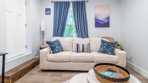 Una sala de estar con un sofá color beige, cojines azules y blancos, una mesa de centro de madera, una cesta de mimbre con revistas, una lámpara de pie blanca y una ventana con cortinas azules.