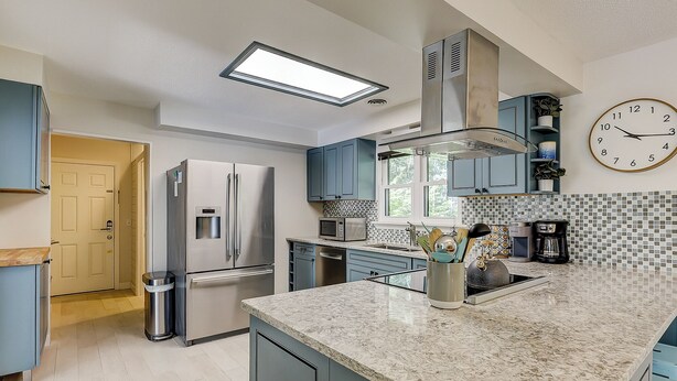 Una cocina moderna con un refrigerador de acero inoxidable, un microondas y una cafetera. Hay una ventana que proporciona luz natural y un reloj en la pared.