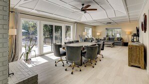 Una habitación amplia con una gran mesa de conferencias rodeada de sillas, un ventilador de techo y vistas a un área exterior a través de puertas correderas grandes.