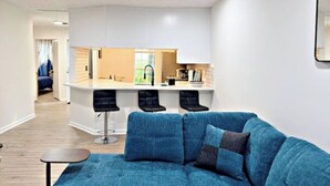Een moderne woonkamer met een blauwe bank, een kleine ronde tafel en een keukenhoek met een bargedeelte en drie barkrukken.