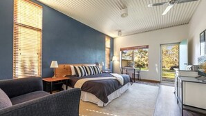 Une chambre avec un lit, un bureau et une chaise. Il y a une fenêtre avec des stores, un ventilateur de plafond et une vue sur l’extérieur.