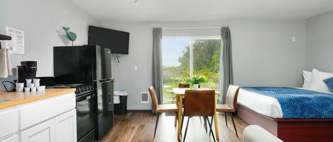 Een moderne keuken met een fornuis en koelkast, een eethoek met een tafel en stoelen, een bed met een blauwe deken en een groot raam met uitzicht op groen.