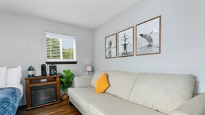 Ein modernes Wohnzimmer mit einem beigefarbenen Sofa, einer blauen Decke auf einem Bett, einem Kamin und drei gerahmten Bildern an der Wand.