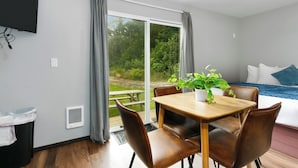 Ein modernes Zimmer mit einem Holzespeisetisch, Ledersesseln, einer Topfpflanze, einem Bett mit blauer und weißer Bettdecke und einem großen Fenster mit Vorhängen.