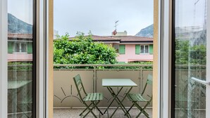 Un balcon avec une table et des chaises, une vue sur un immeuble aux volets verts et une haie.