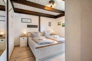Un dormitorio con techo de madera, cama con ropa de cama blanca, mesita de noche con lámpara y pared con cuadros enmarcados.