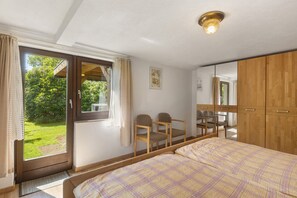 Una camera da letto con un letto, un'armadio in legno, una scrivania con sedie, una finestra con tende e vista su del verde all'esterno.