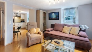 Un salon avec un canapé, un fauteuil et une table basse, une cuisine à l’arrière-plan et une fenêtre donnant sur un paysage urbain.