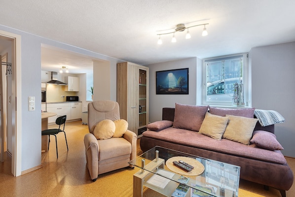 Un salón con sofá, sillón y mesa de centro, una cocina al fondo, y un ventanal con vista panorámica de la ciudad.