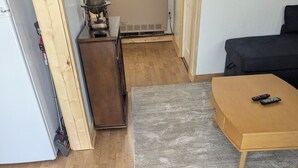 A room with a wooden floor, a grey rug, a wooden table with two remotes, a small cabinet, and a radiator.