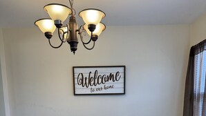 A dining room with a wooden table, white chairs, a chandelier, and a welcome sign on the wall. - Main Street Condo in Miles City (Miles City)