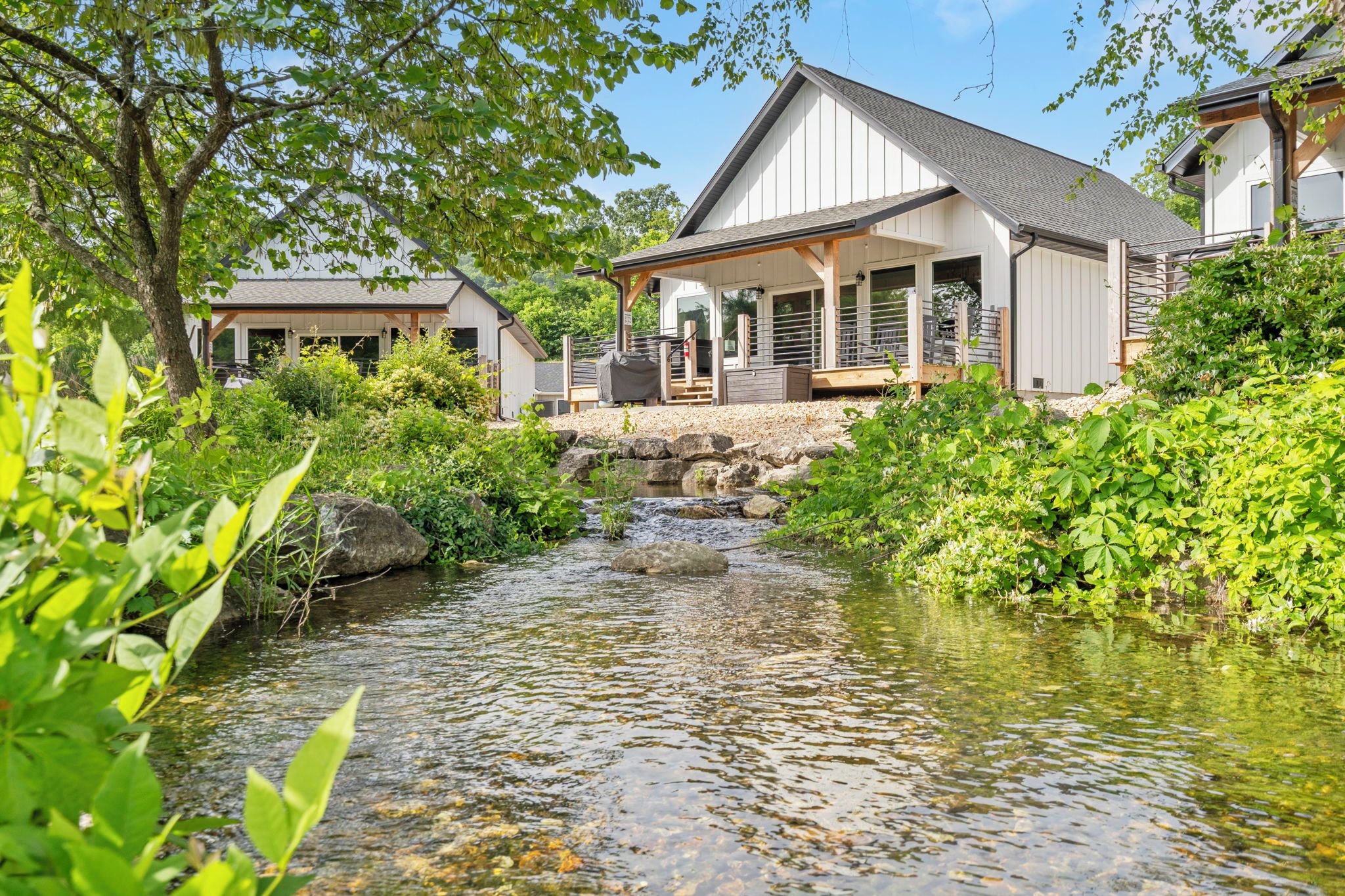 Ein Haus mit einer überdachten Veranda, einem kleinen Bach davor und einem Garten mit verschiedenen Pflanzen.