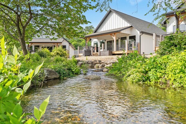 A house with a covered porch, a small stream in front, and a garden with various plants.