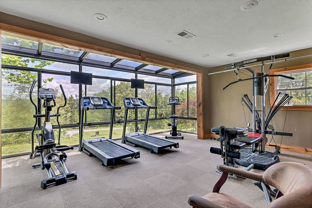 A gym with treadmills, weight machines, and a rowing machine, large windows, and a view of trees.