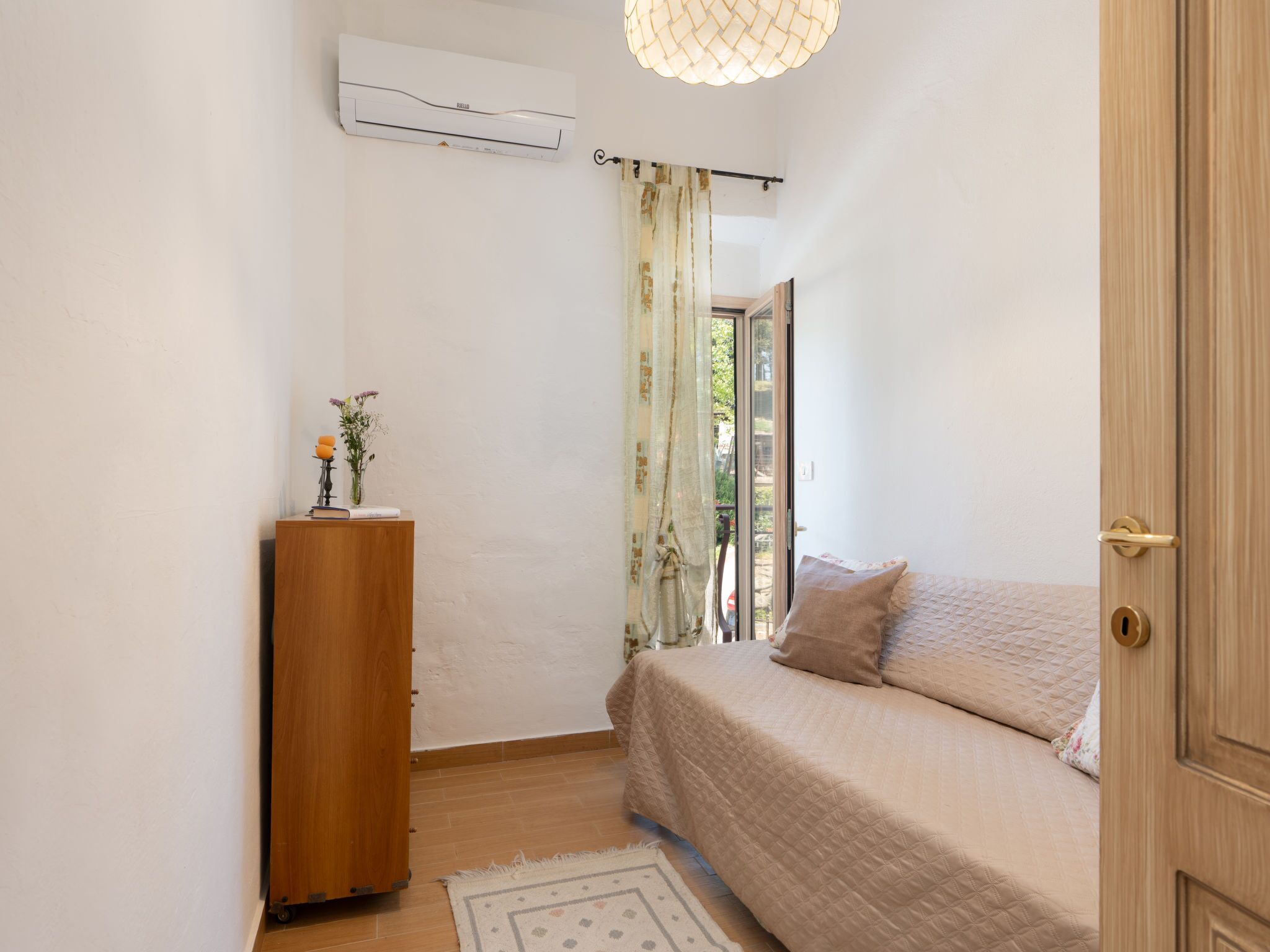 Un dormitorio con cama, cómoda de madera, aire acondicionado y vista a través de una ventana con cortinas.
