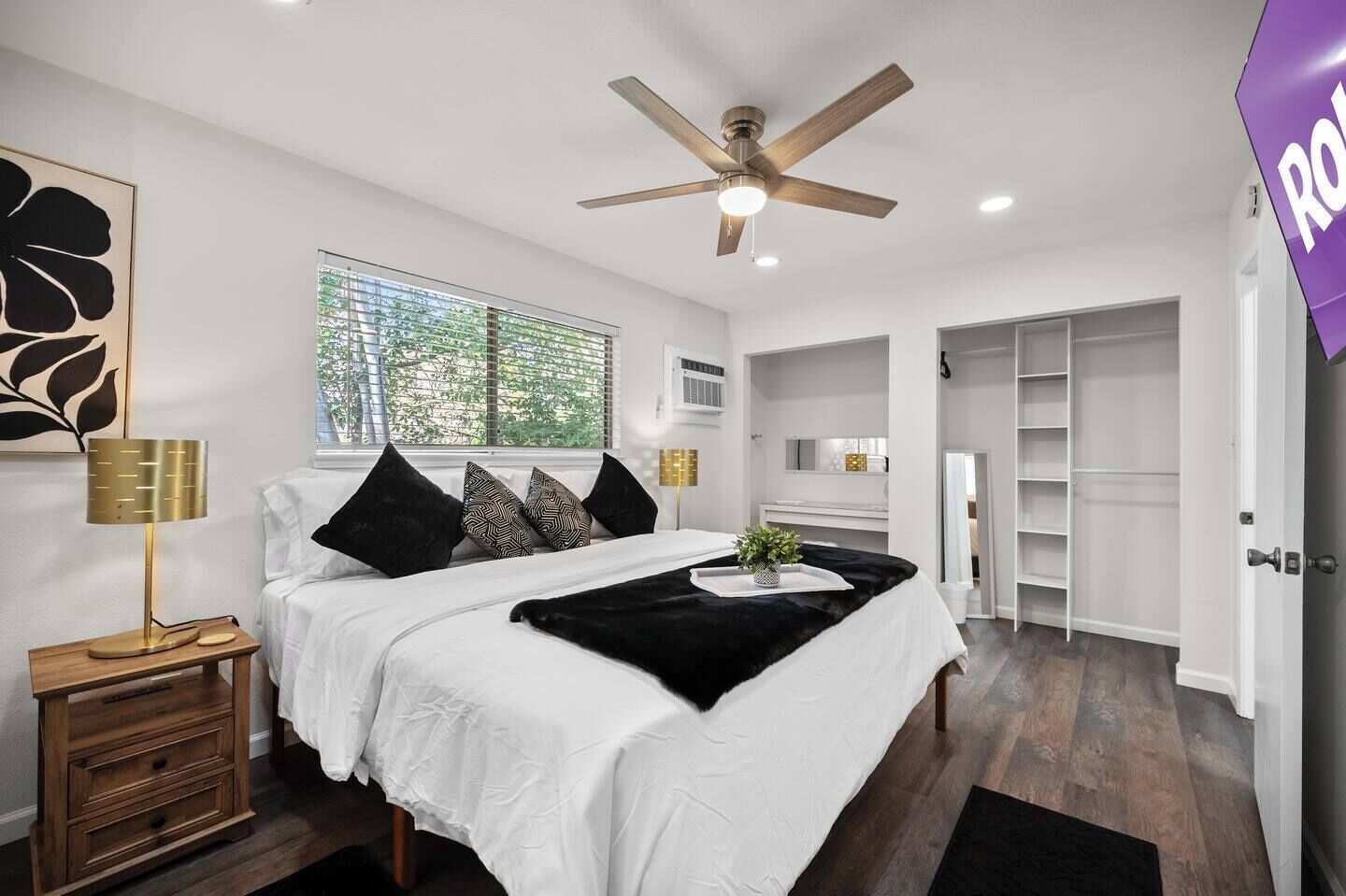 Quarto moderno com uma cama grande, um ventilador de teto, uma janela com persianas, uma mesinha de cabeceira com abajur e uma televisão na parede.