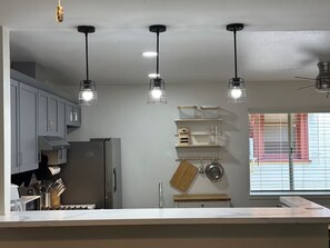 Een moderne keuken met grijze kastjes, een roestvrijstalen koelkast en hangende lampen. Er is een raam met zonwering en een plank met keukengerei.