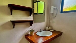 Une salle de bain avec un meuble-lavabo en bois, un lavabo blanc, un miroir, une petite plante en pot, des toilettes et une fenêtre donnant sur un paysage verdoyant.